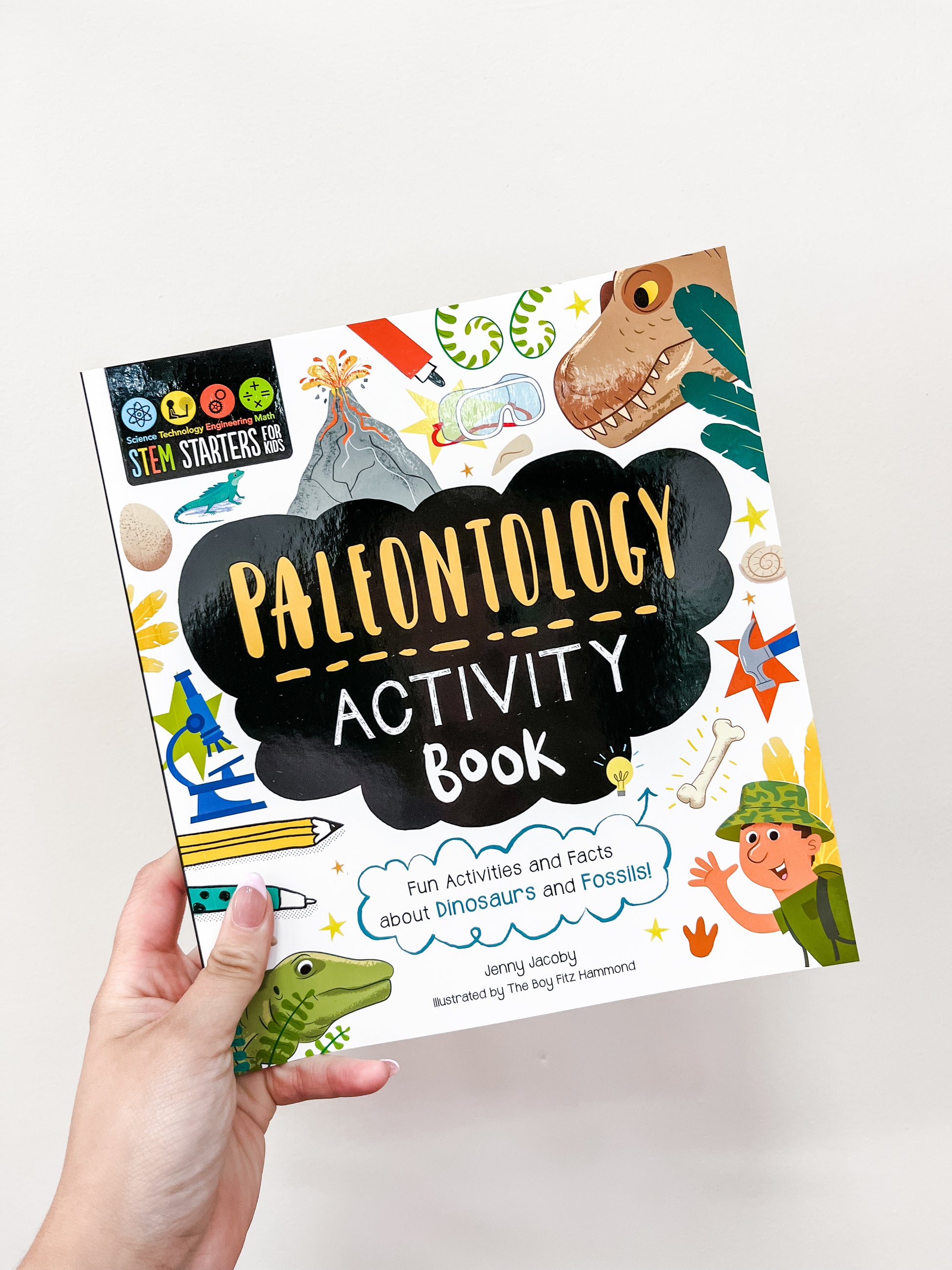 A hand holds the "Stem Starters Art Activity Book: Paleontology" by Simon & Schuster, with a colorful cover of cartoon dinosaurs and fossils, offering fun activities and facts about paleontology.