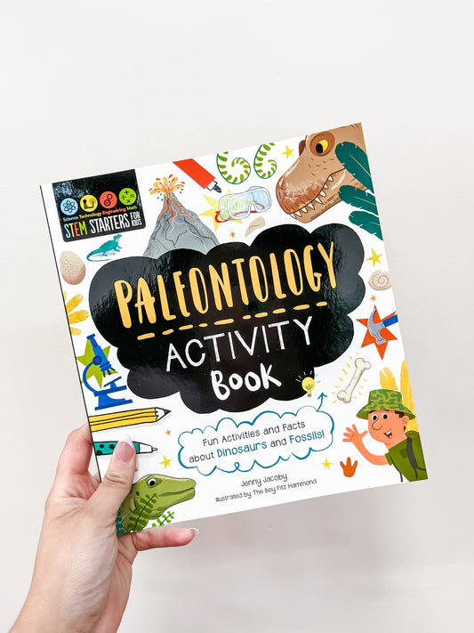 A hand holds the "Stem Starters Art Activity Book: Paleontology" by Simon & Schuster, with a colorful cover of cartoon dinosaurs and fossils, offering fun activities and facts about paleontology.