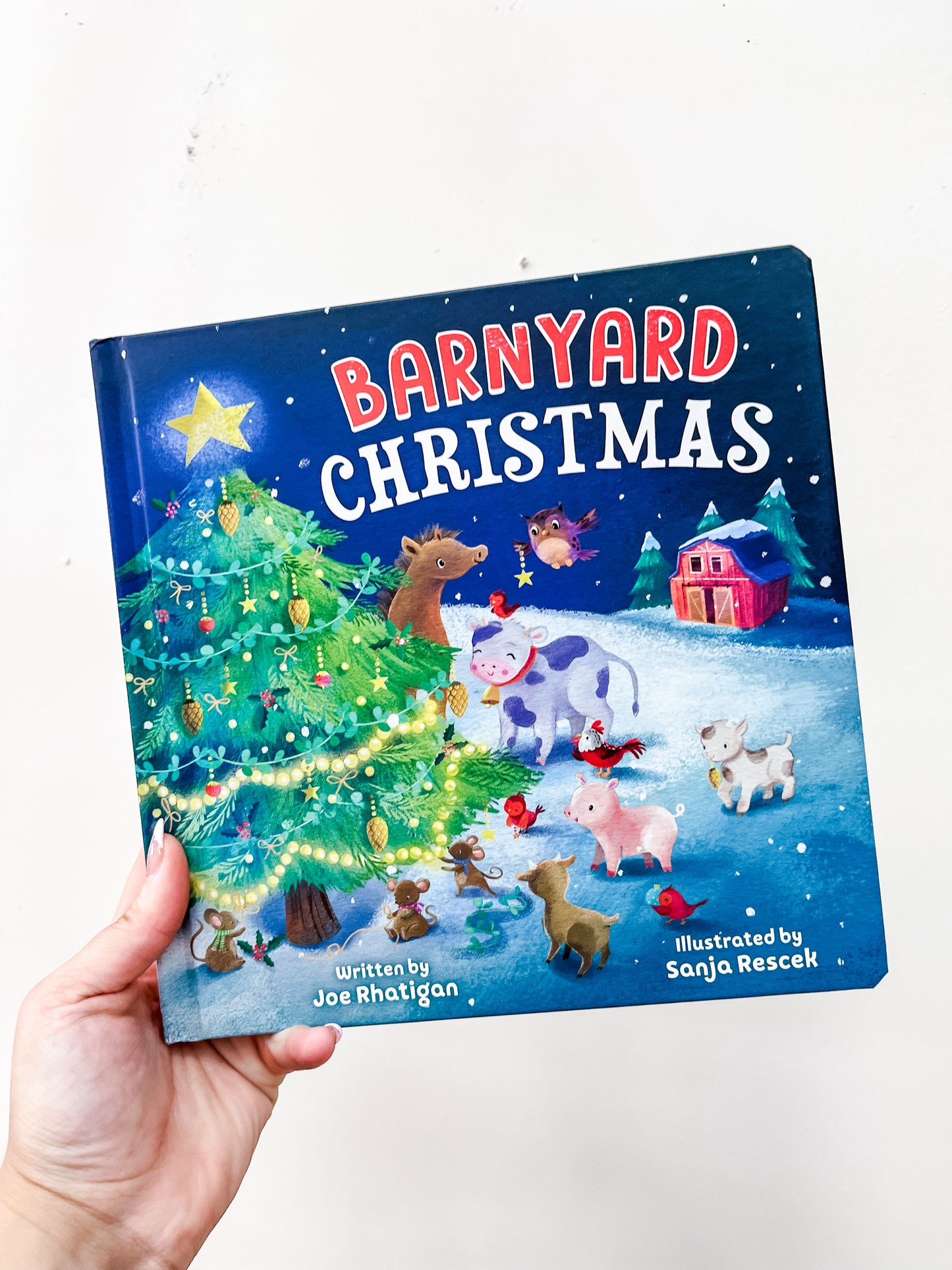 A hand holds the Barnyard Christmas Book by Simon & Schuster, featuring a decorated Christmas tree, farm animals, a red barn, and snow on the cover.