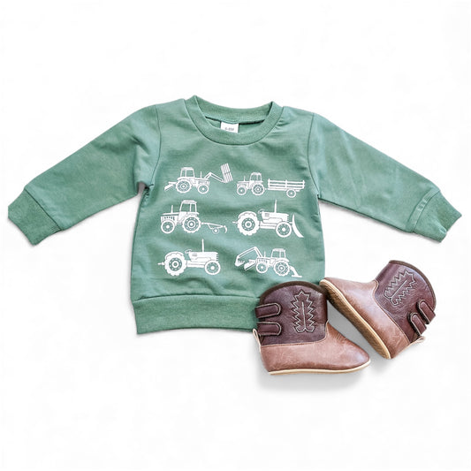 The Little Farmer Pullover by Tiny Tot Bundles, a green children’s sweatshirt with white tractor prints, is paired with small brown cowboy boots on a white background.