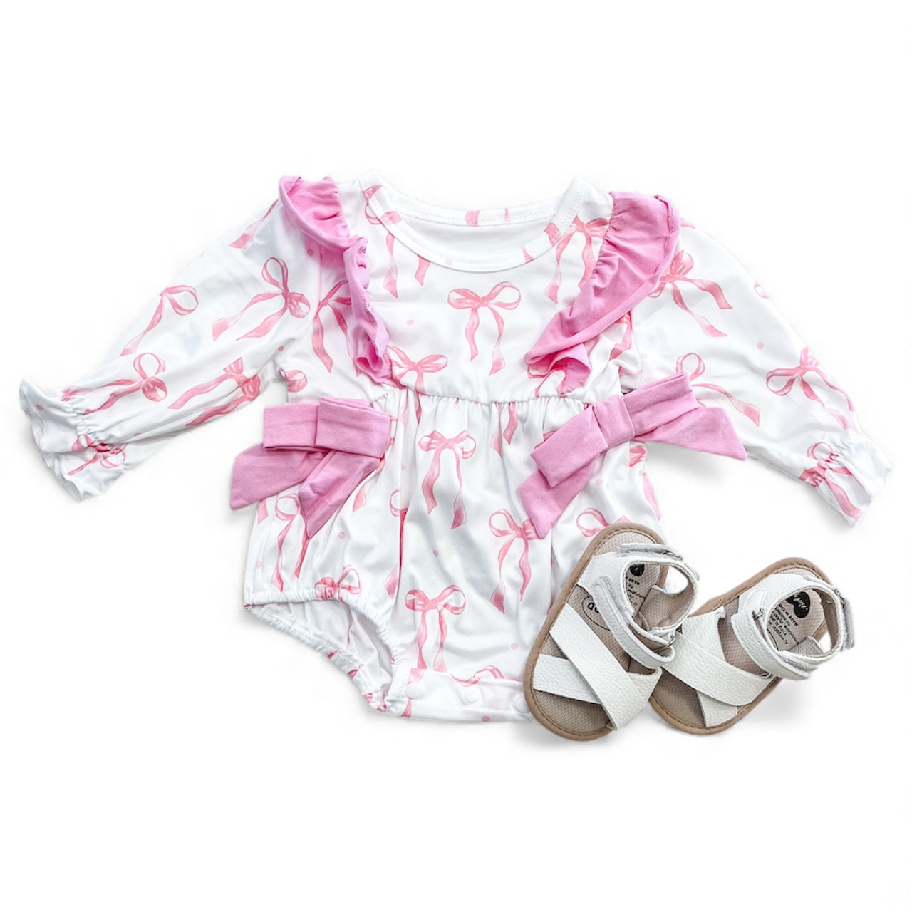 The Ribbon Pink Cozy Romper by Tiny Tot Bundles features a white long-sleeve design with pink ribbon patterns and ruffle accents, paired with white baby sandals with tan soles.