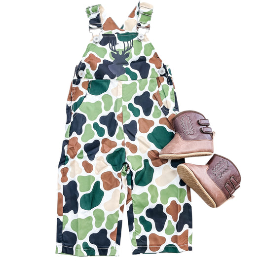 The Little Hunter Overalls by Tiny Tot Bundles, featuring a black deer silhouette on camouflage fabric, are paired with brown cowboy boots with white stitching, shown against a white background.