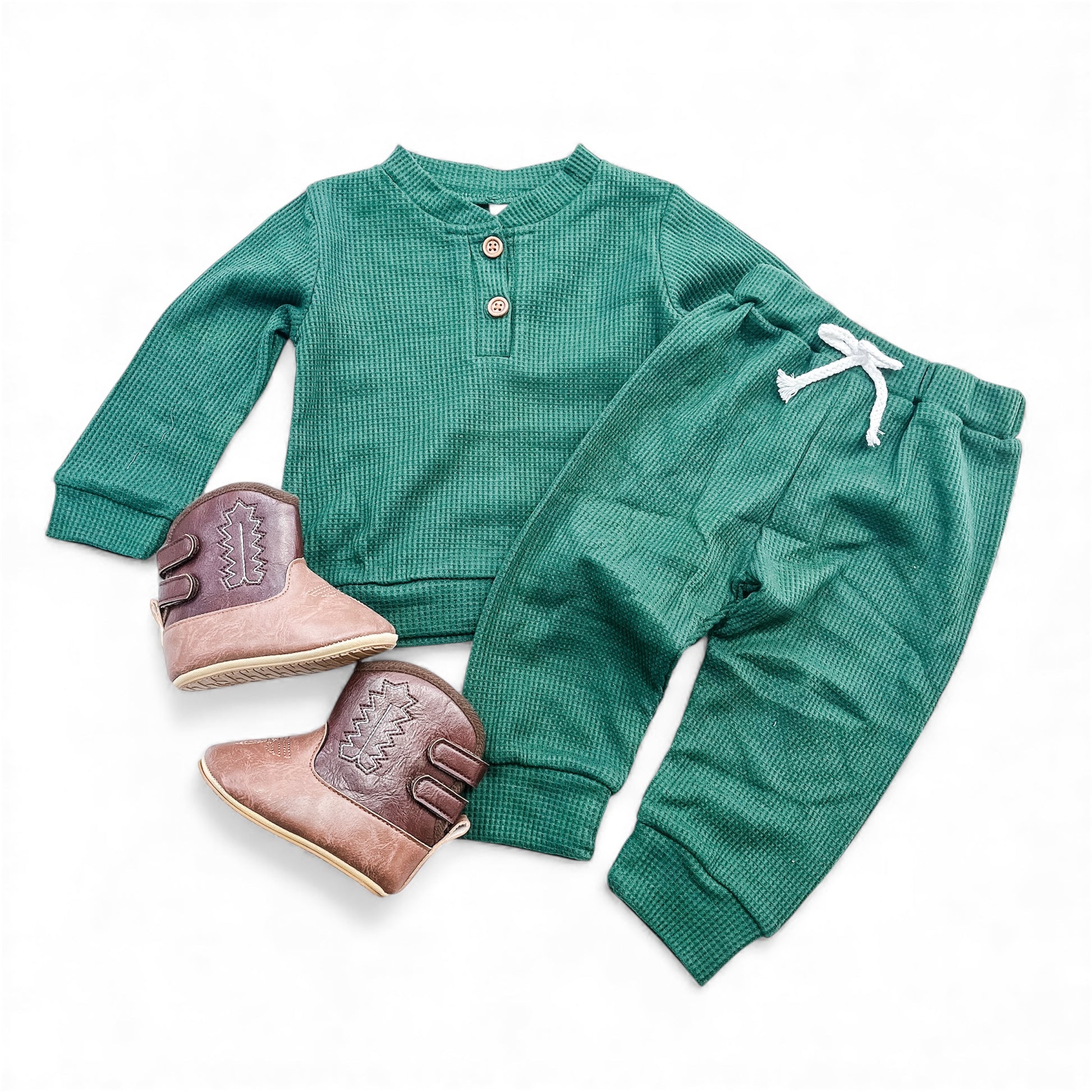 The Tiny Tot Bundles Everyday Waffle Knit Set, featuring a green long-sleeve buttoned top and matching pants with a white drawstring, is paired with brown ankle boots with decorative stitching, displayed on a white background.
