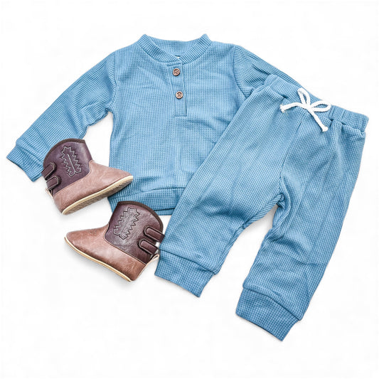 The Tiny Tot Bundles Everyday Waffle Knit Set, a blue ribbed long-sleeve shirt and matching pants for toddlers, is shown next to brown and tan cowboy boots on a white background.