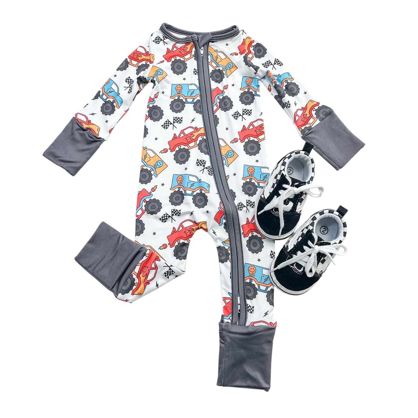 The Tiny Tot Bundles "Monster Rally Zippy," a baby onesie featuring a colorful monster truck print with gray cuffs and front zipper, is paired with black and white baby sneakers, displayed on a white background.