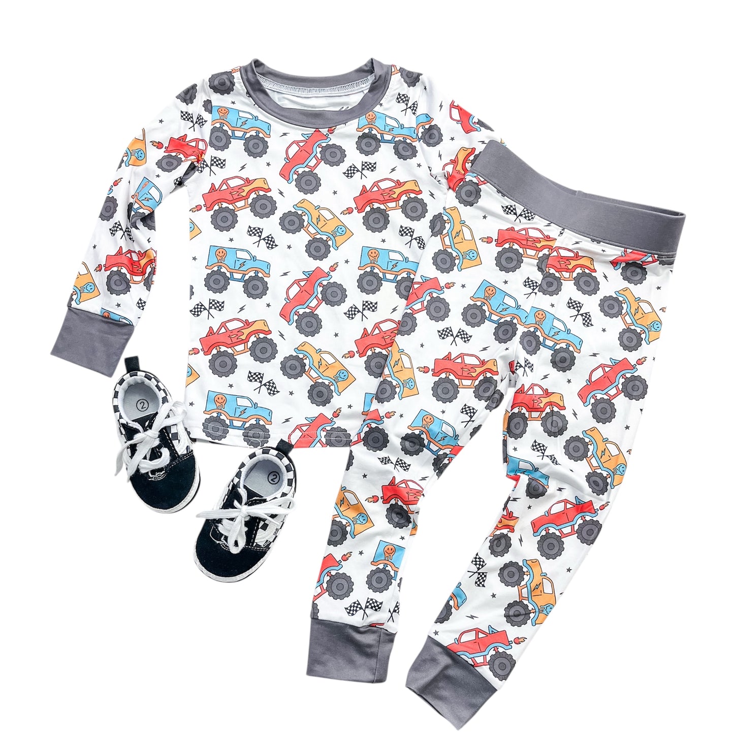 The Tiny Tot Bundles Monster Rally Jammies Set features colorful monster truck patterns, a long-sleeve top and pants with gray cuffs, shown paired with black and white sneakers.