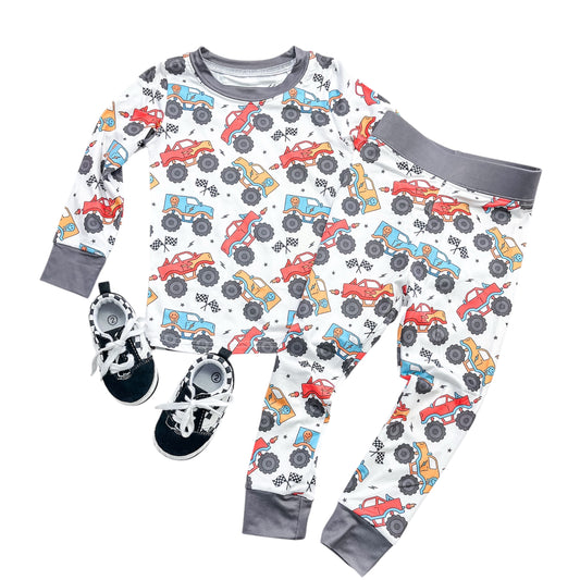 The Tiny Tot Bundles Monster Rally Jammies Set features colorful monster truck patterns, a long-sleeve top and pants with gray cuffs, shown paired with black and white sneakers.