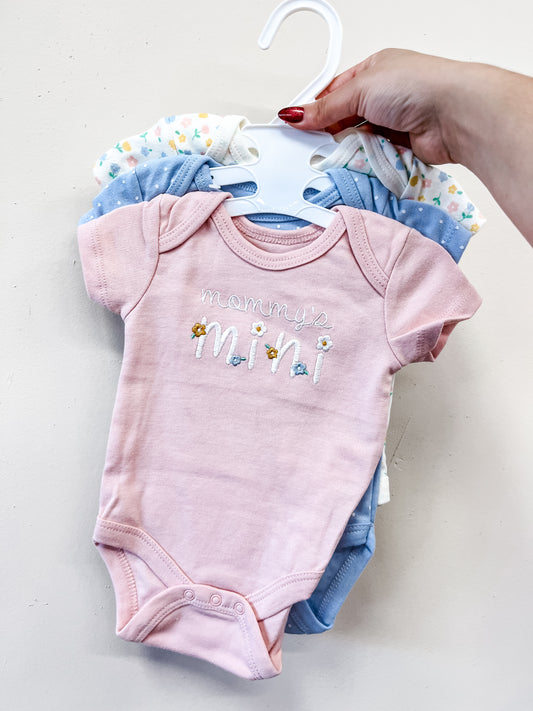 A hand holds a hanger with three Lollipop Dreams Mommy’s Mini Bubble Trio bodysuits: the front pink one reads "mommy’s mini" with decorative designs, while blue and white ones with subtle patterns are behind it.