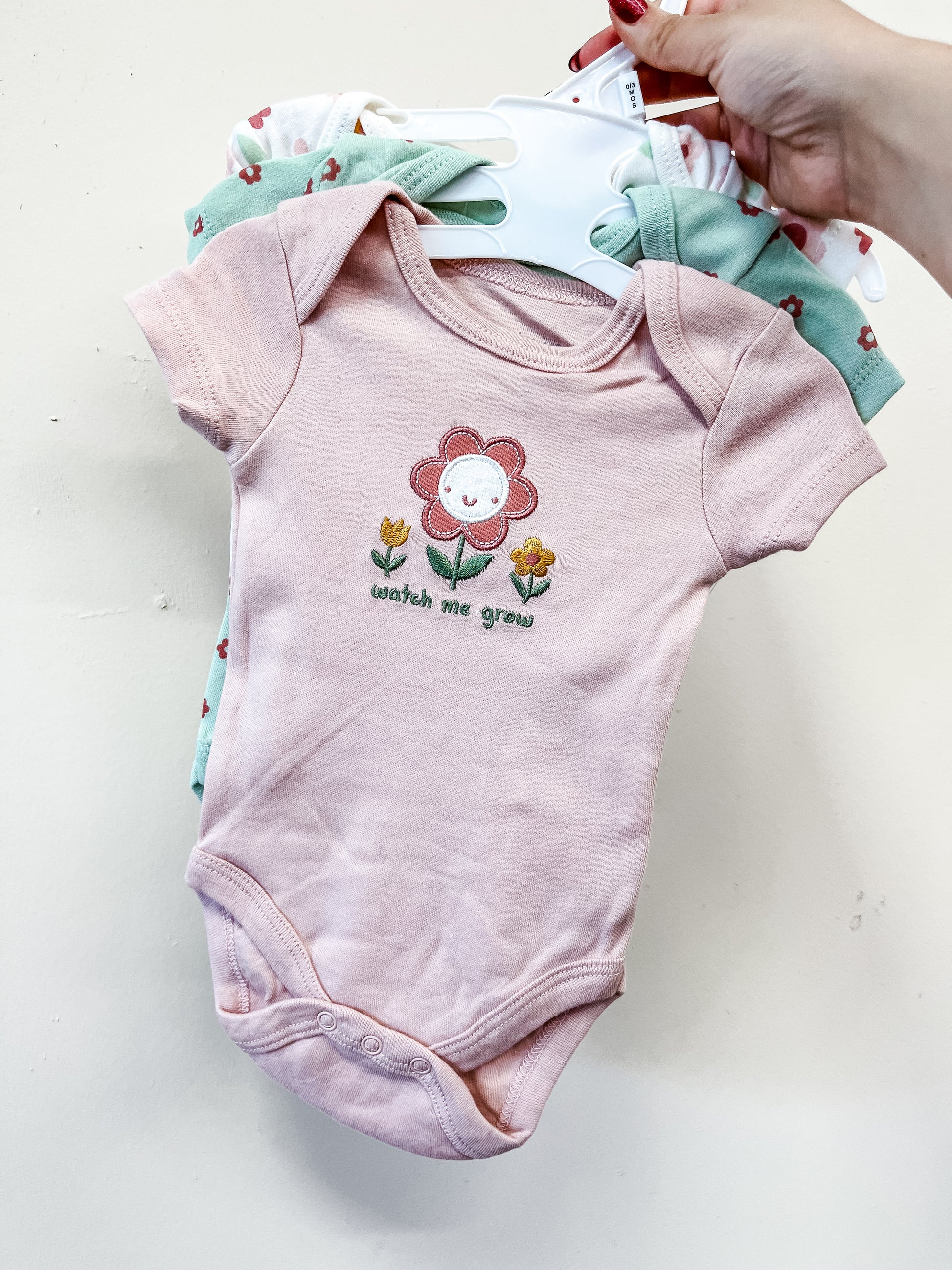 A hand holds up a set of Lollipop Dreams Flower Power Bubble Trio baby onesies on hangers; the front is pink with a smiling flower and "watch me grow" printed on it, while other pastel onesies are visible behind.