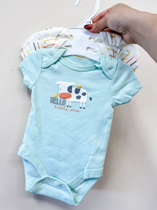 A hand holds a hanger with Lollipop Dreams' Tiny Farm Bubble Trio. The front onesie is light blue with a cow and chick graphic and "Hello little one." Other colorful striped onesies are partially visible behind it.