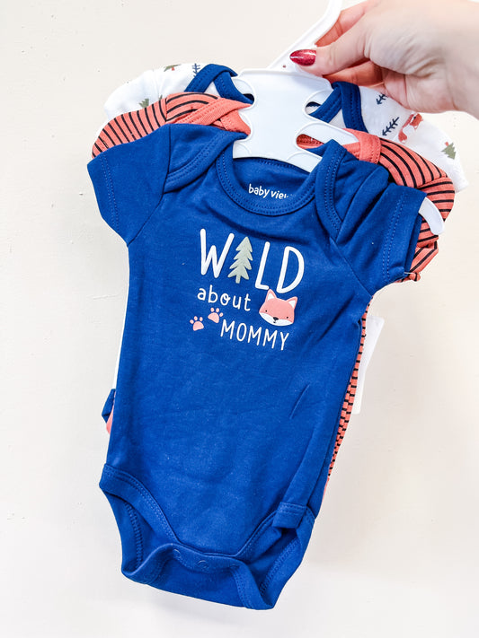 A hand holds the Lollipop Dreams Wild About Mommy Bubble Trio onesies on hangers. The front blue onesie features a fox and "Wild about Mommy" text, with other patterned onesies from the set partially visible behind it.