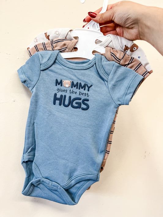 A hand holds up three "Mommy Hugs Bubble Trio" onesies by Lollipop Dreams—one light blue with “Mommy gives the best hugs” and a bear face, and two beige with animal prints.
