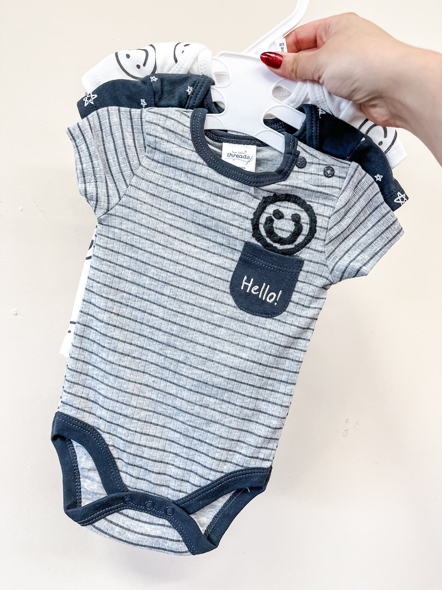 A hand holds Lollipop Dreams’ Hello Happy Bubble Trio onesies on hangers. The front gray striped onesie features navy trim, a “Hello!” pocket, and a smiley face; similar designs from the set are seen behind it.