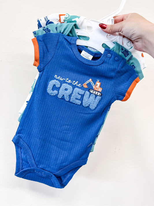 A hand holds a set of Lollipop Dreams Tiny Crew Bubble Trio baby onesies on hangers. The front blue onesie with orange trim features construction vehicles and the phrase "new to the CREW" on the chest.
