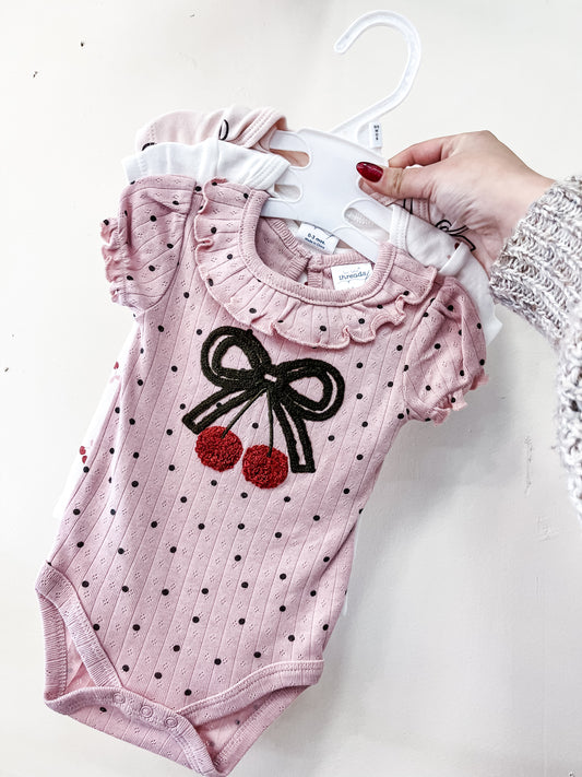 A hand holds three Lollipop Dreams Cherry Bow Bubble Trio baby onesies on hangers; the front is pink with black polka dots, frilled sleeves, and a bow topped with two embroidered cherries.