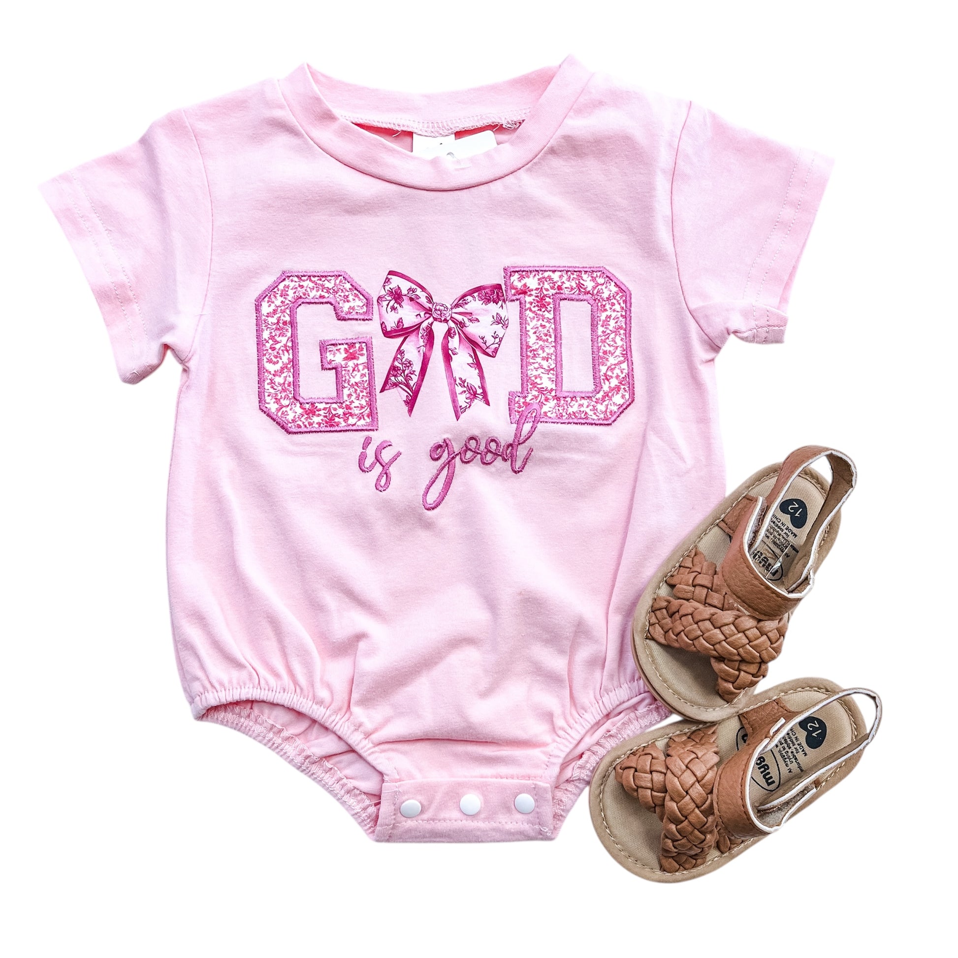 The Tiny Tot Bundles God Is Good Bow Bubble, a pink baby onesie with sparkly lettering and bow design, is paired with brown woven sandals and displayed on a white background.