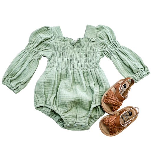 The Willow Green Smocked Bubble by Tiny Tot Bundles, a long-sleeved baby romper, is paired with brown woven toddler sandals placed to the right of the outfit on a white background.
