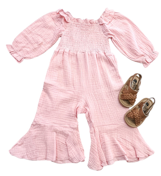 The Blush Meadow Smocked Romper by Tiny Tot Bundles, a pink long-sleeve ruffled toddler jumpsuit with flared legs, is laid flat next to a pair of brown woven toddler sandals.