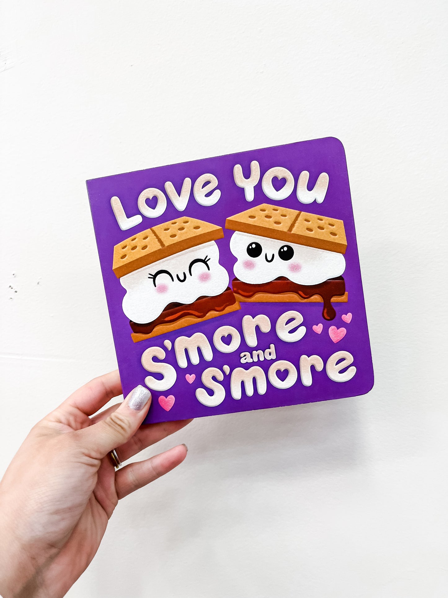 A hand displays the "Love You S'more and S'more" card by Simon & Schuster, featuring two smiling kawaii s’mores with blushing cheeks and pink hearts—perfect for lovers of cute food-themed puns.
