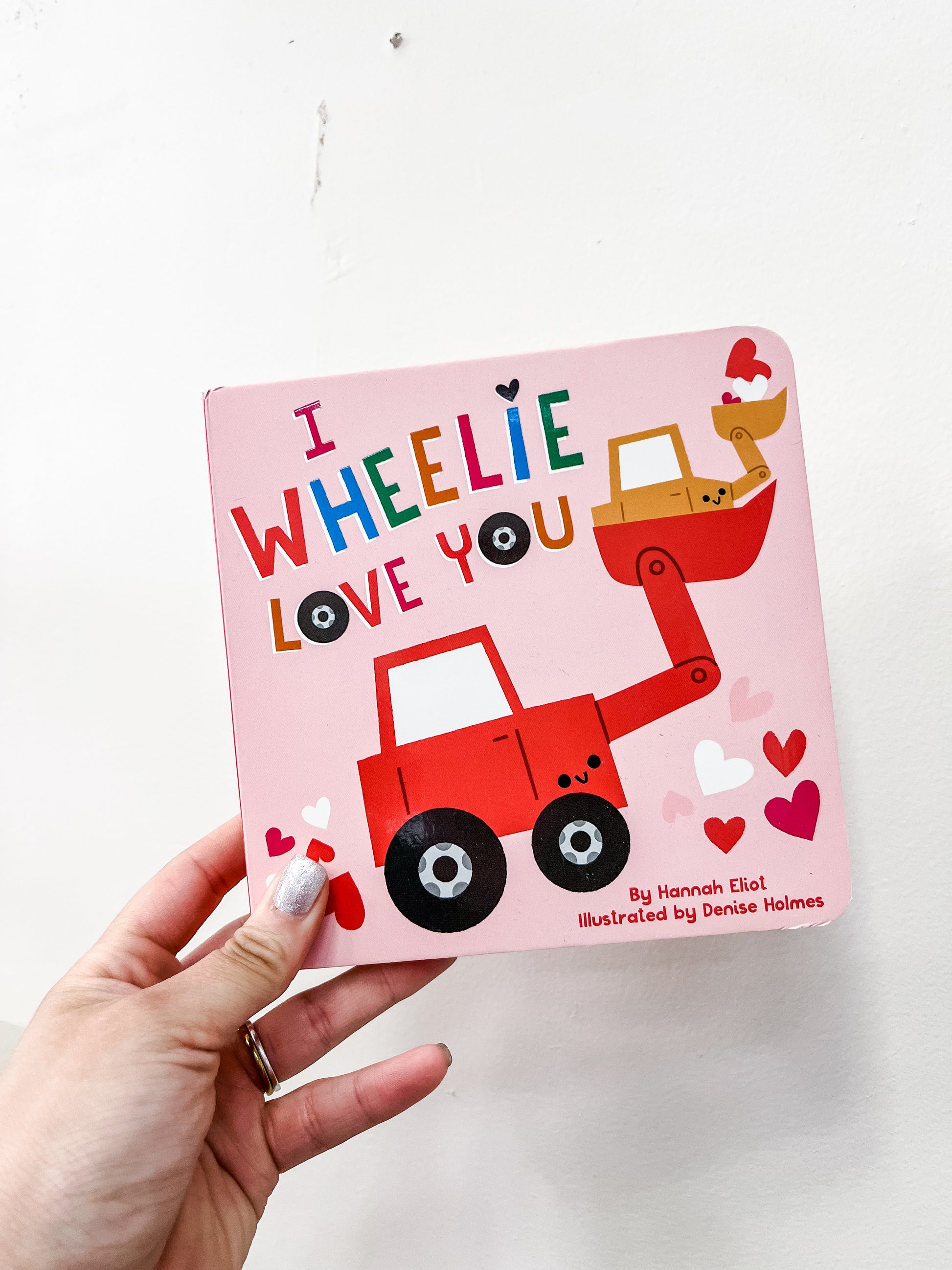 A hand holds the “I Wheelie Love You Book” by Simon & Schuster, a pink Valentine’s Day board book featuring a red and yellow cartoon excavator with hearts on the cover.