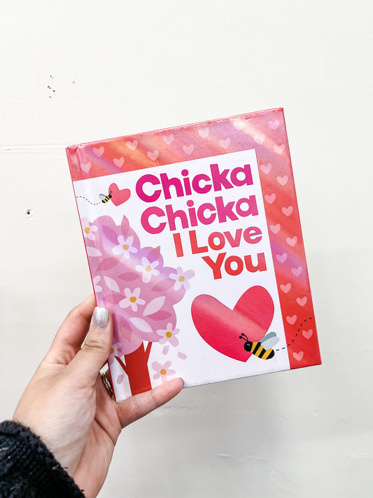 A hand holds the Chicka Chicka I Love You Book by Simon & Schuster, featuring a pink and red cover with a heart, flowers, bees, and a tree—an adorable Valentine’s Day gift. The background is a plain white wall.