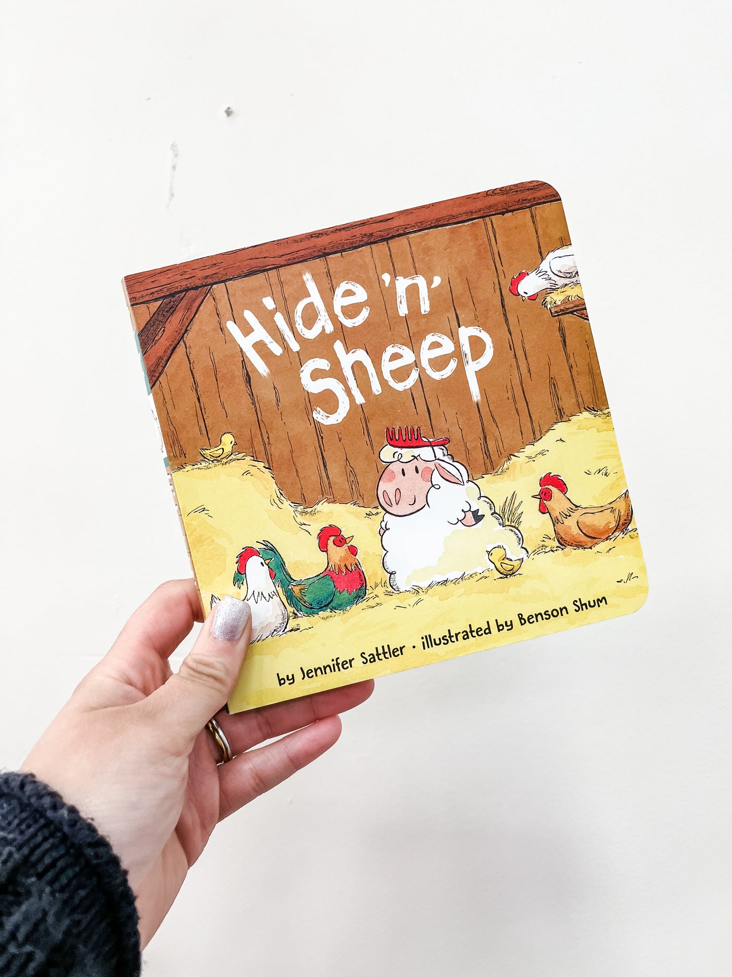 A hand holds the Hide 'n' Sheep Book by Simon & Schuster. The cover features a sheep with glasses sitting in hay among chickens, with a hen house in the background.