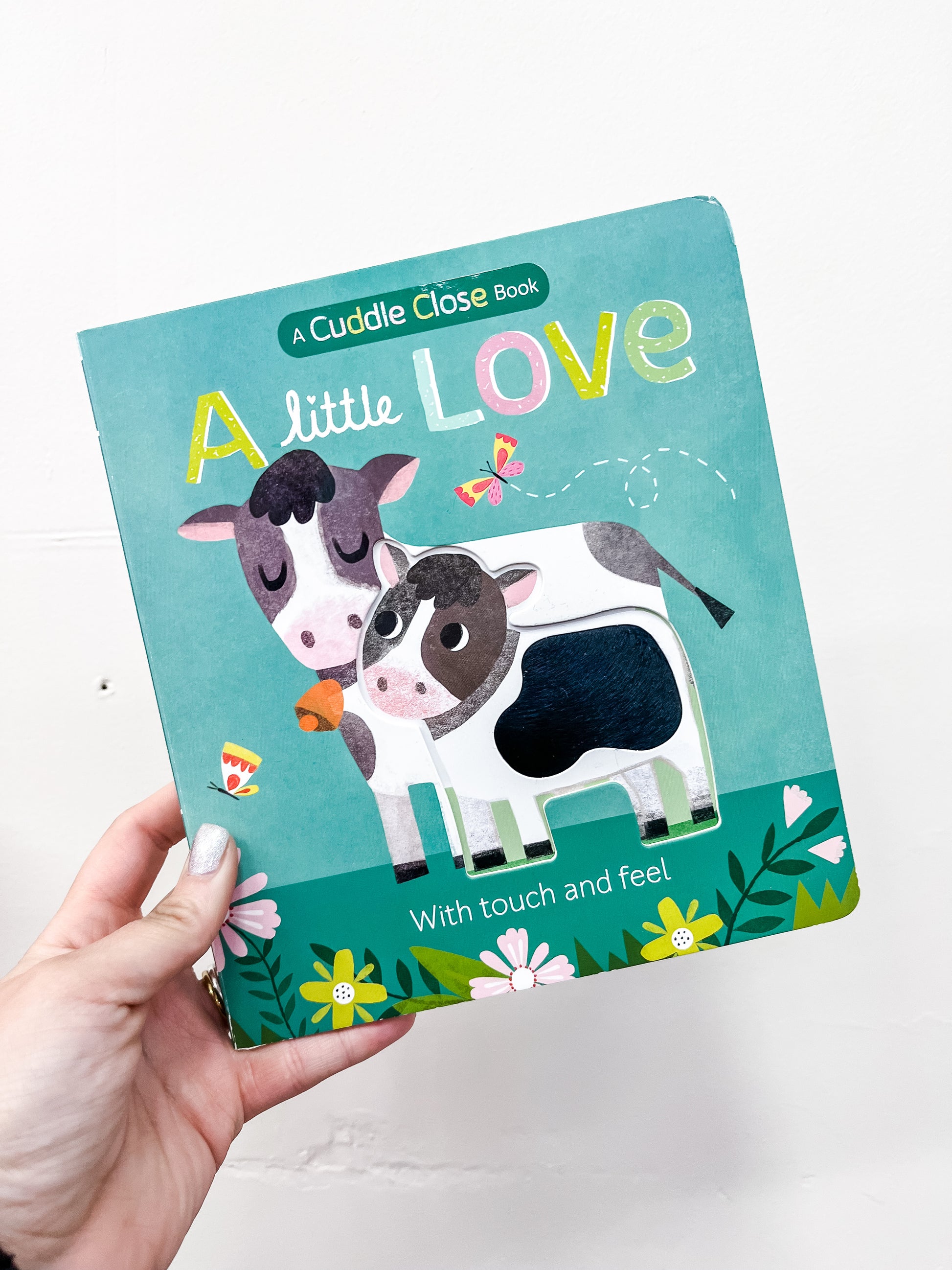 A hand holds Simon & Schuster’s Little Love Book, a touch-and-feel board book for kids, featuring two cartoon cows with butterflies and flowers on the green cover—perfect for little hands.