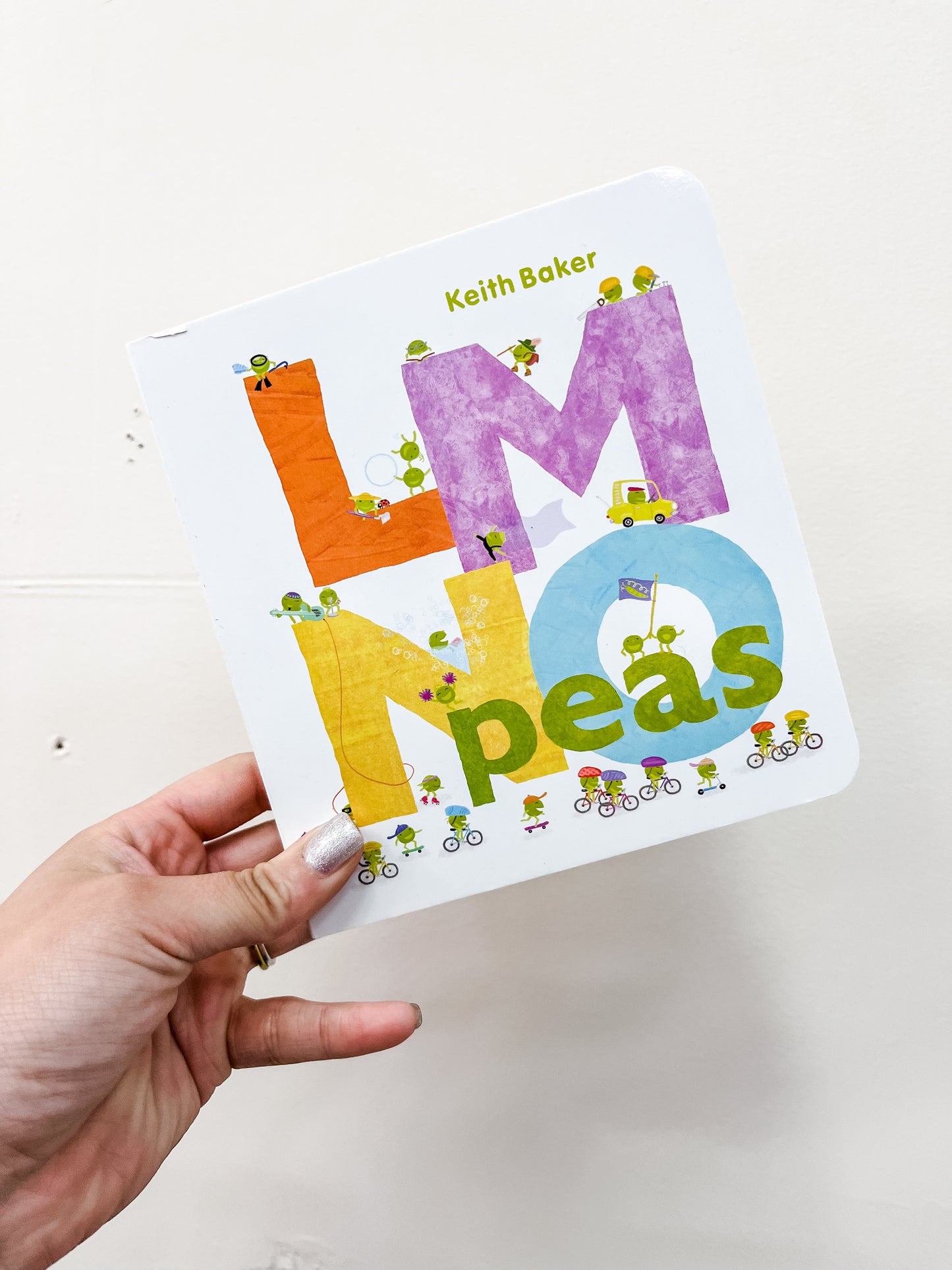 A hand with silver nail polish holds the Lmno Peas Book by simon, showcasing large colorful letters and tiny illustrated peas doing different activities on the cover.