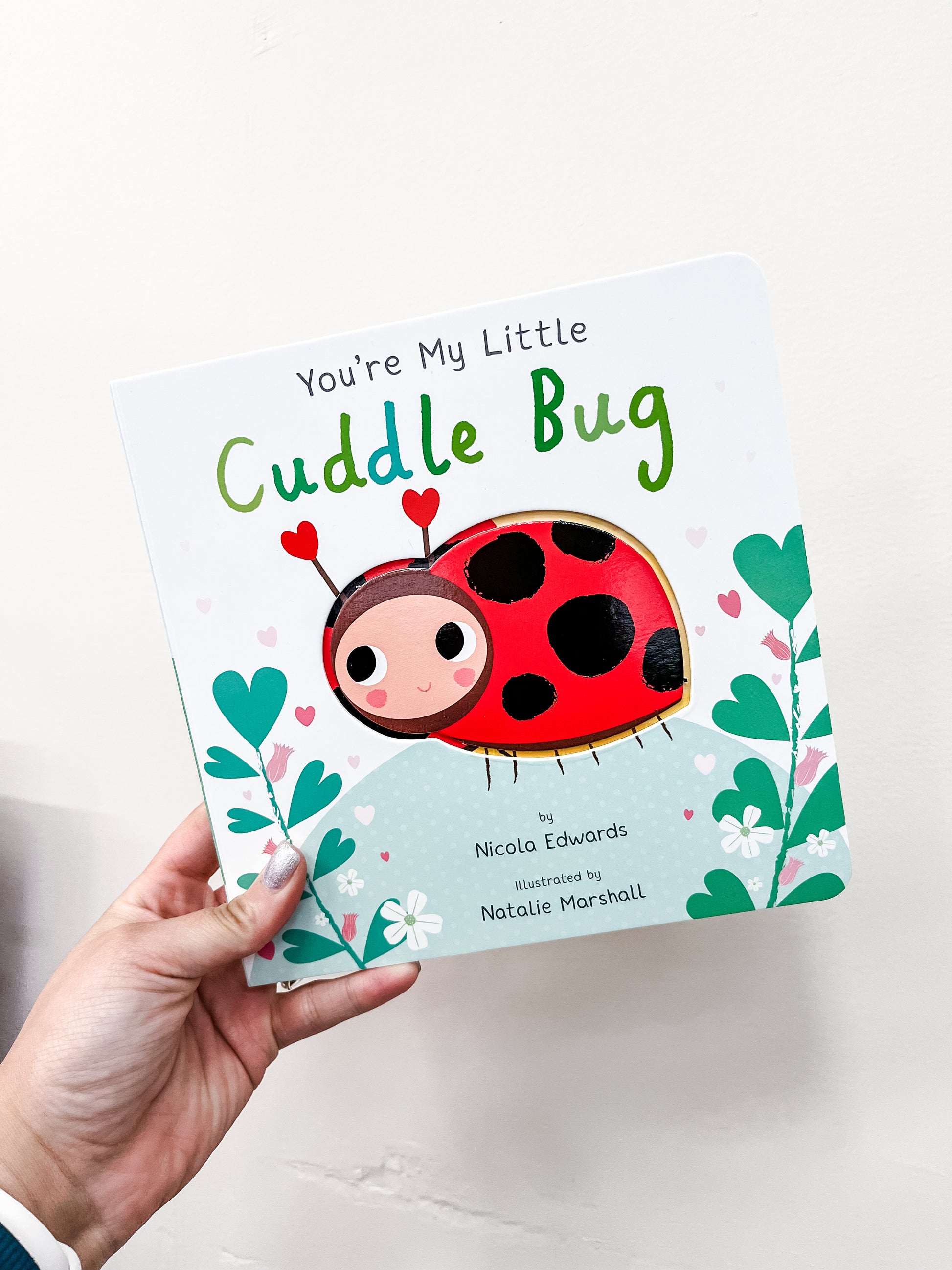 A hand holds the board book "You're My Little Cuddle Bug" from Simon & Schuster, featuring a smiling ladybug with heart-shaped antennae on the chunky cover.