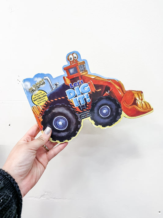 A hand holds the "Let's Dig It Book" by Simon & Schuster, shaped like an orange construction vehicle, against a plain white wall.