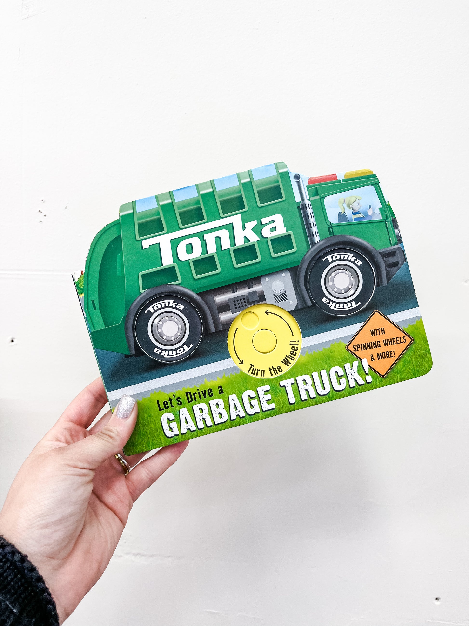 A hand holds the Tonka: Let's Drive A Garbage Truck Book by Simon & Schuster, an interactive board book shaped like a green garbage truck with a cover featuring a yellow button labeled “Turn the wheel!”.