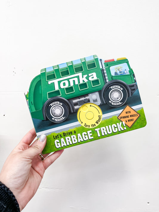 A hand holds the Tonka: Let's Drive A Garbage Truck Book by Simon & Schuster, an interactive board book shaped like a green garbage truck with a cover featuring a yellow button labeled “Turn the wheel!”.