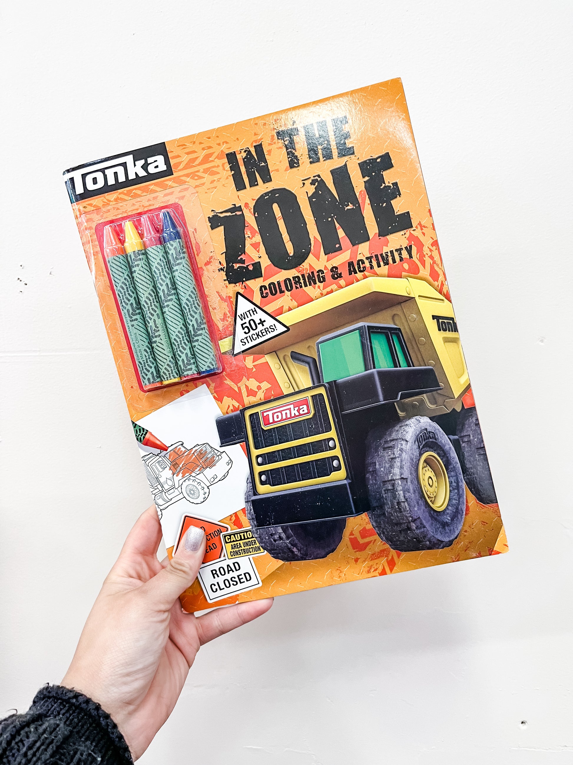 A hand holds Simon & Schuster's Tonka: in the Zone: Coloring & Activity Book, featuring a yellow dump truck on the cover, plus crayons and stickers—ideal for kids’ mazes, puzzles, and coloring fun.