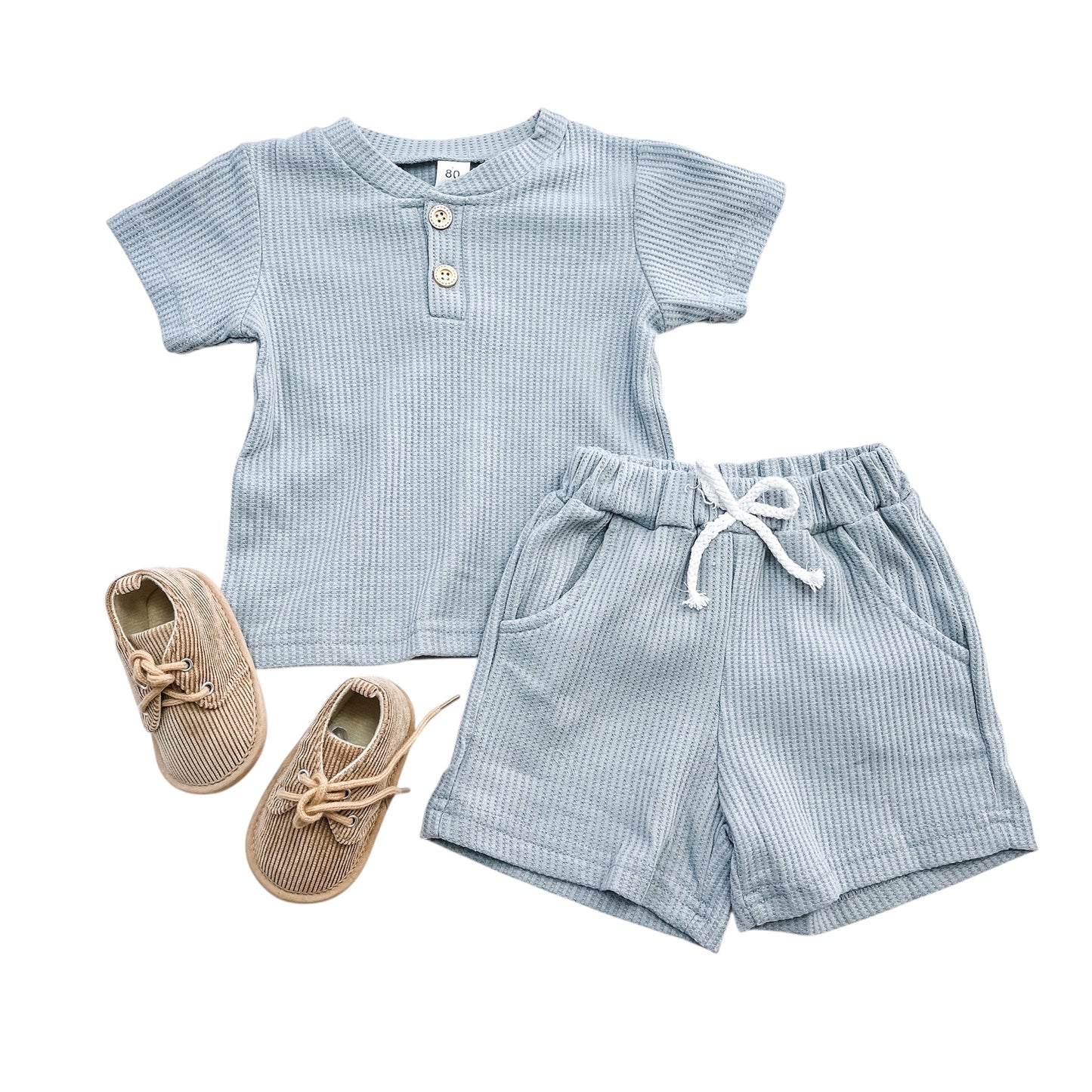 The Essentials Boys Set by Tiny Tot Bundles, featuring a light blue short-sleeve Henley shirt with buttons, matching drawstring shorts, and beige lace-up baby shoes, is laid out on a white background.