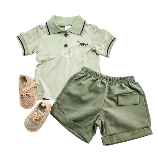 The Little Dino Polo Set by Tiny Tot Bundles features a light green short-sleeve polo with a dinosaur graphic, matching green shorts with a back pocket, and tan lace-up baby shoes, all displayed on a white background.