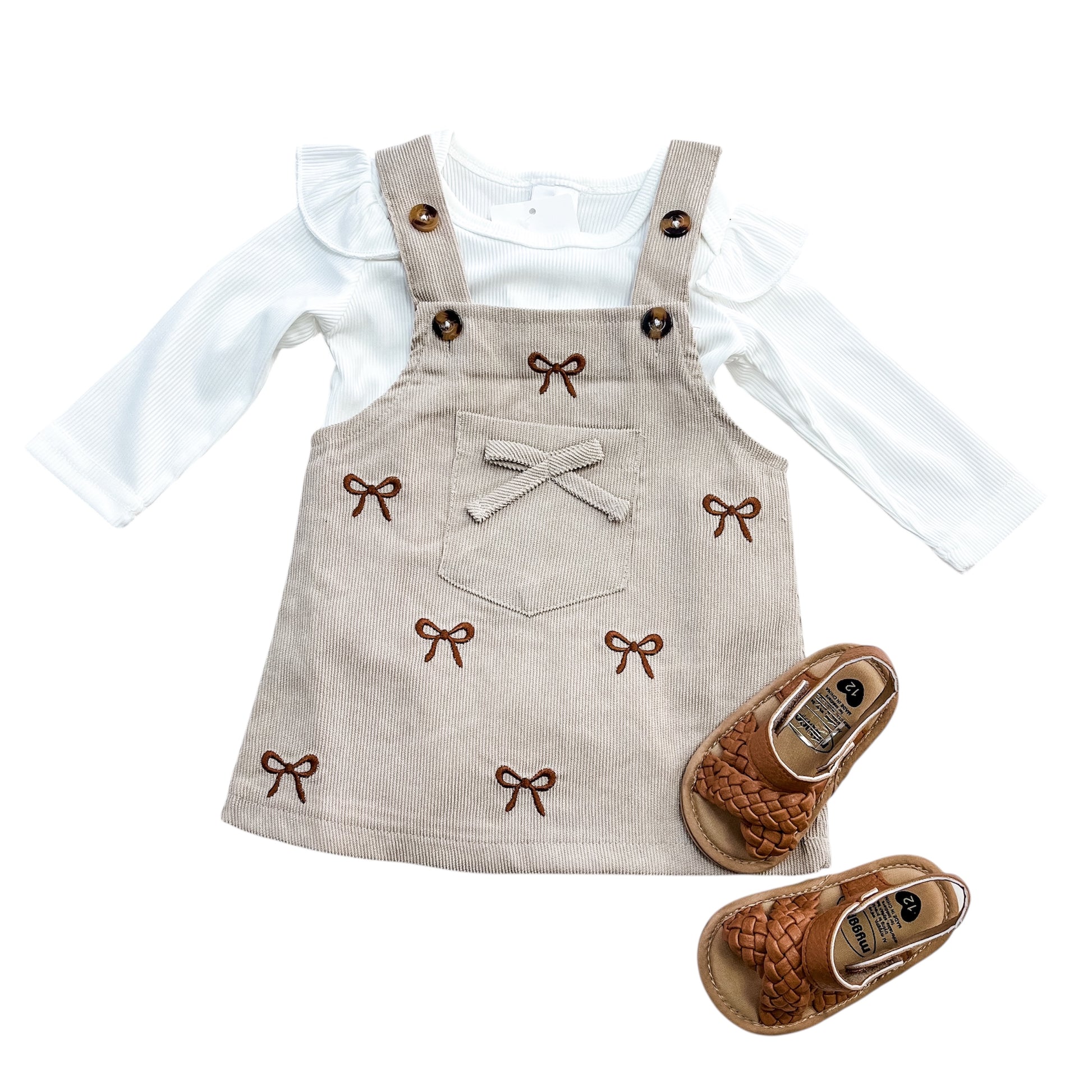 The Tiny Tot Bundles Corduroy Coquette Suspender Set features a white ruffle-sleeve top, a beige corduroy pinafore with brown bow details and a front pocket, paired with brown woven baby sandals.