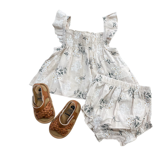 The Flutter Bloom Baby Set by Tiny Tot Bundles features a cream floral ruffled top, matching bloomers, and tan woven sandals, all shown against a white background.