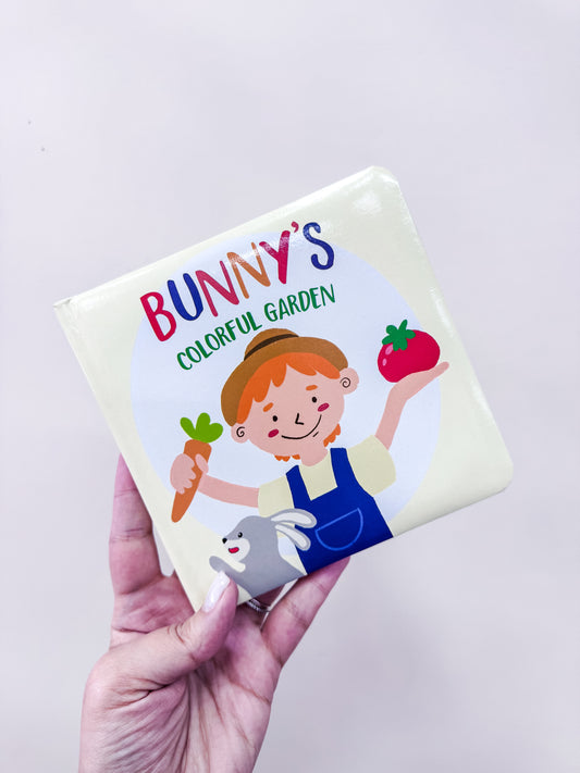 A hand holds the "Colorful Vegetable Garden Board Book" by Tiny Tot Bundles, featuring a child with a carrot and strawberry, and a smiling bunny at the bottom.