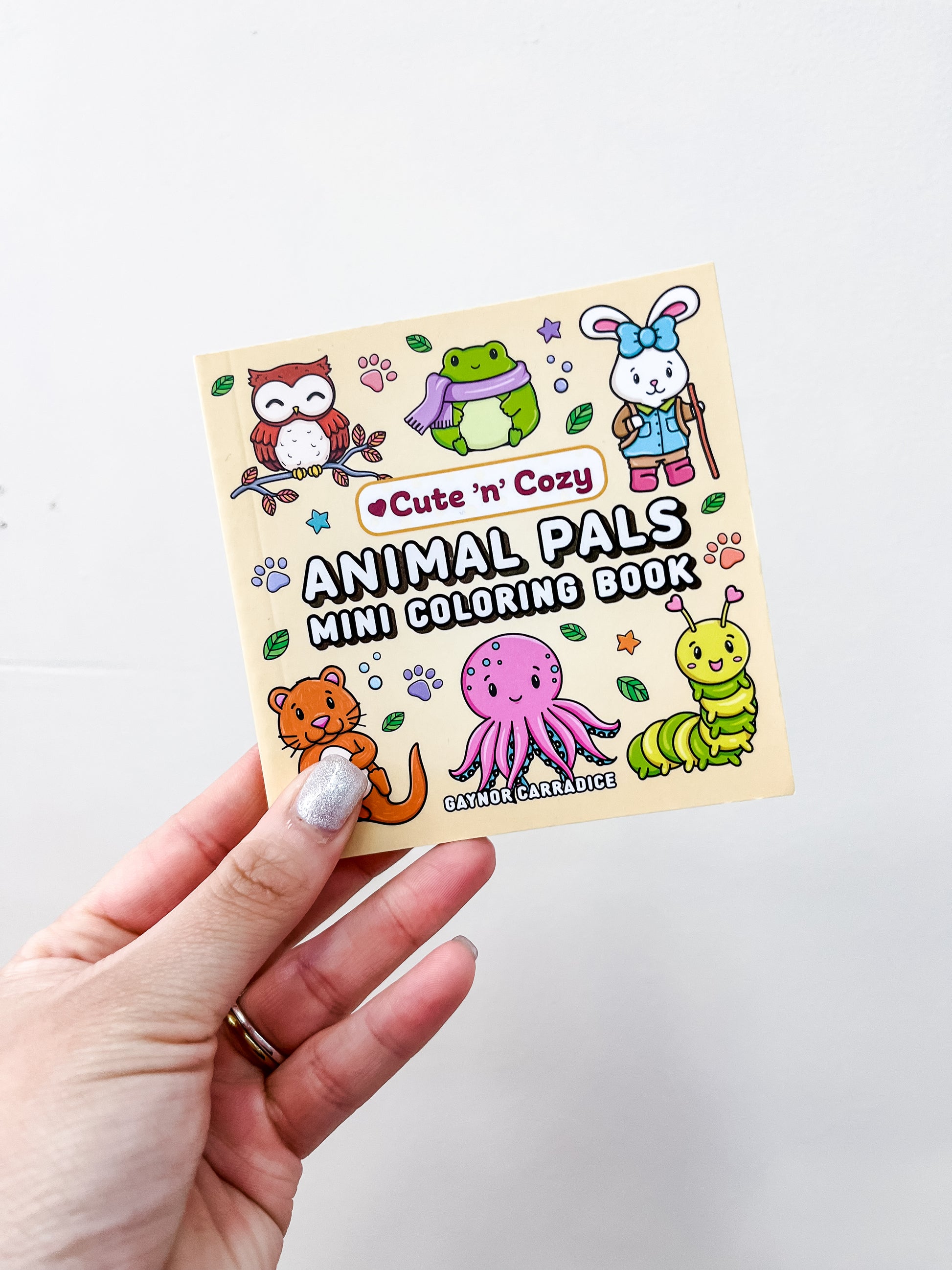 A hand holds the Animal Pals Mini Coloring Book by Simon & Schuster, featuring cozy animal designs like an owl, frog, rabbit, cat, octopus, and caterpillar on the cover.