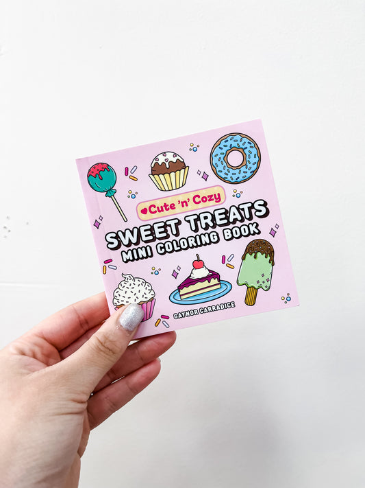 A hand with silver nail polish holds the Simon & Schuster Sweet Treats Mini Coloring Book—a portable book featuring cupcakes, donuts, cake, and ice cream on the cover.