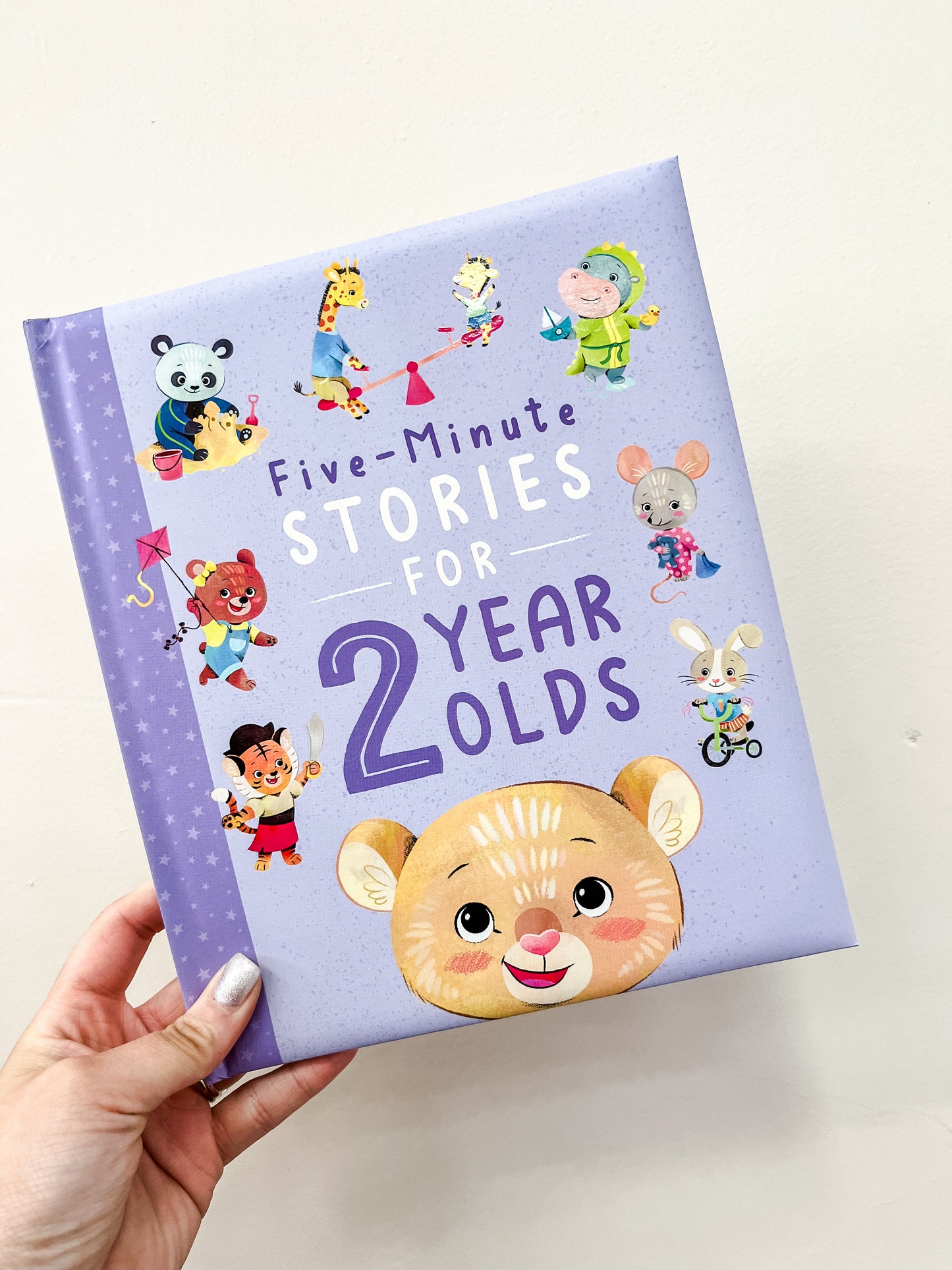 A hand holds the Simon & Schuster Five-Minute Stories For 2 Year Olds Book, featuring a purple cover with cute animal and child illustrations including a bear, panda, mouse, rabbit, and giraffe.