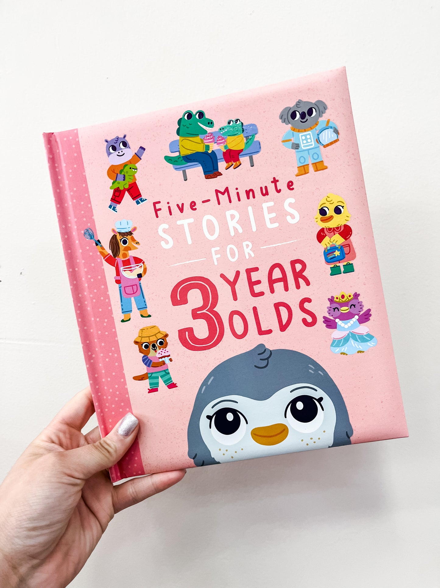 A hand holds the Simon & Schuster Five-Minute Stories For 3 Year Olds Book, a pink children’s book featuring cartoon animals like a penguin, cat, frog, koala, and more on the cover.