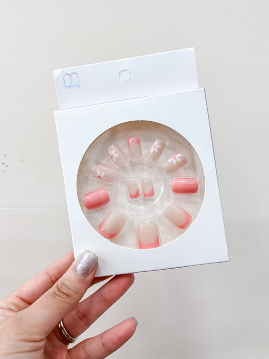 A hand with glittery silver nail polish holds a box of love & repeat’s Daisy Theme Press On Nail Set, featuring pink French tips and white floral designs arranged in a circle inside the packaging.