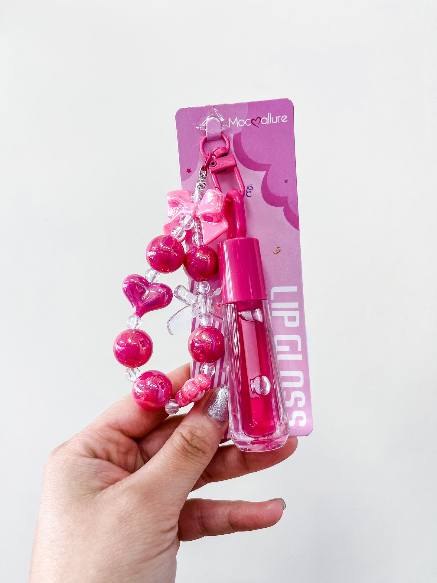 A hand holds a package of love & repeat Gloss & Go Charm Lip Gloss, featuring an attached beaded keychain with pink beads, heart shapes, and a pink bow against a plain white background.