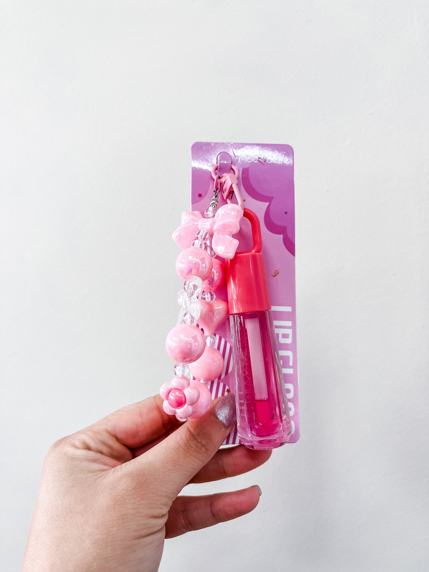 A hand holds love & repeat’s Gloss & Go Charm Lip Gloss in pastel purple and white packaging, decorated with a pink beaded keychain charm, set against a plain white background.
