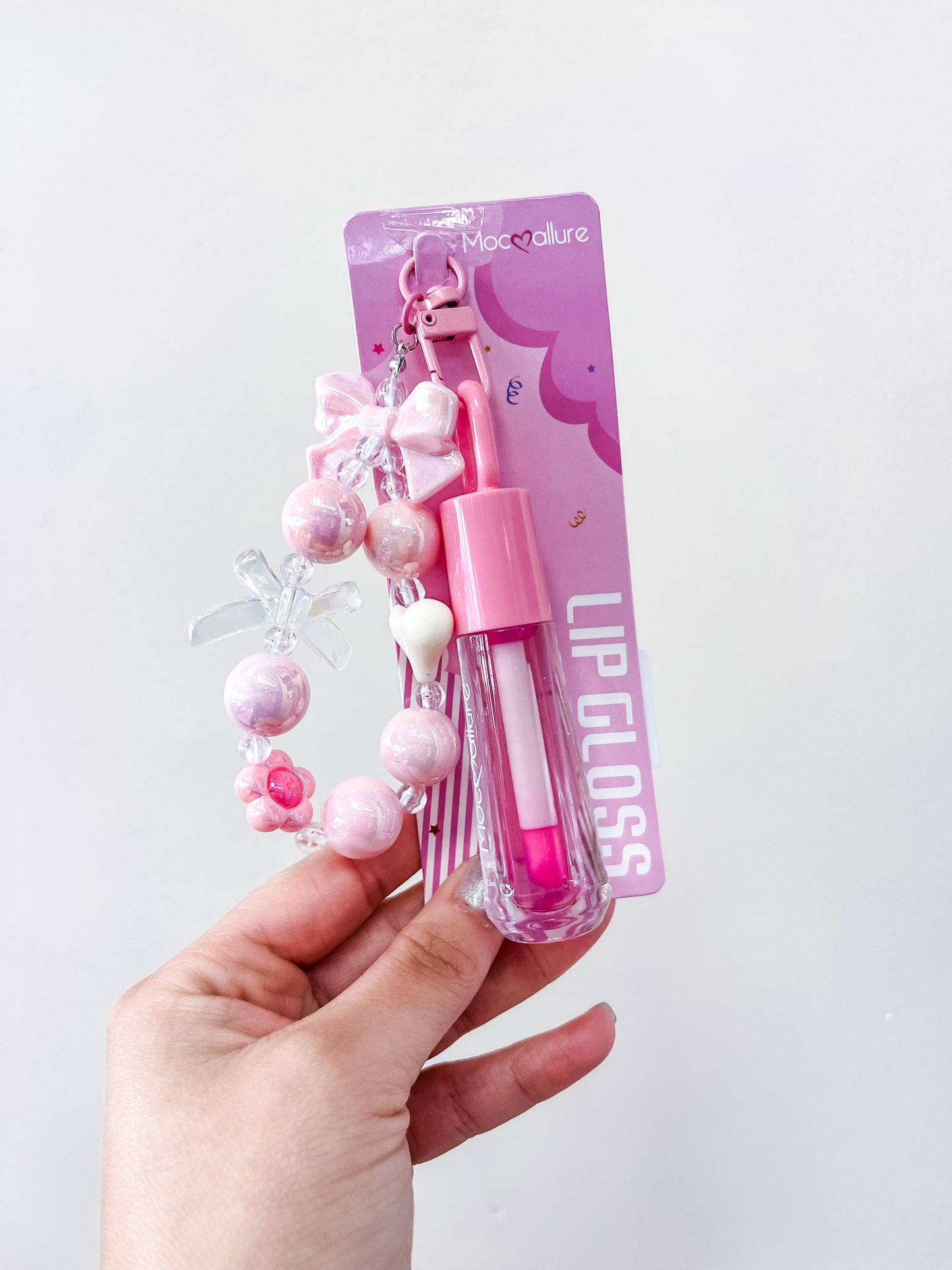 A hand holds a packaged Gloss & Go Charm Lip Gloss from love & repeat, featuring purple and pink packaging labeled "LIP GLOSS" and a matching pastel beaded keychain with small charm decorations.