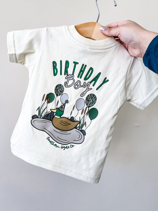 The cream-colored Birthday Boy Balloon T-Shirt by Southern Hyde Co hangs on a wooden hanger, featuring a duck with party hat, balloons, reeds, and "BIRTHDAY BOY" text printed on it.