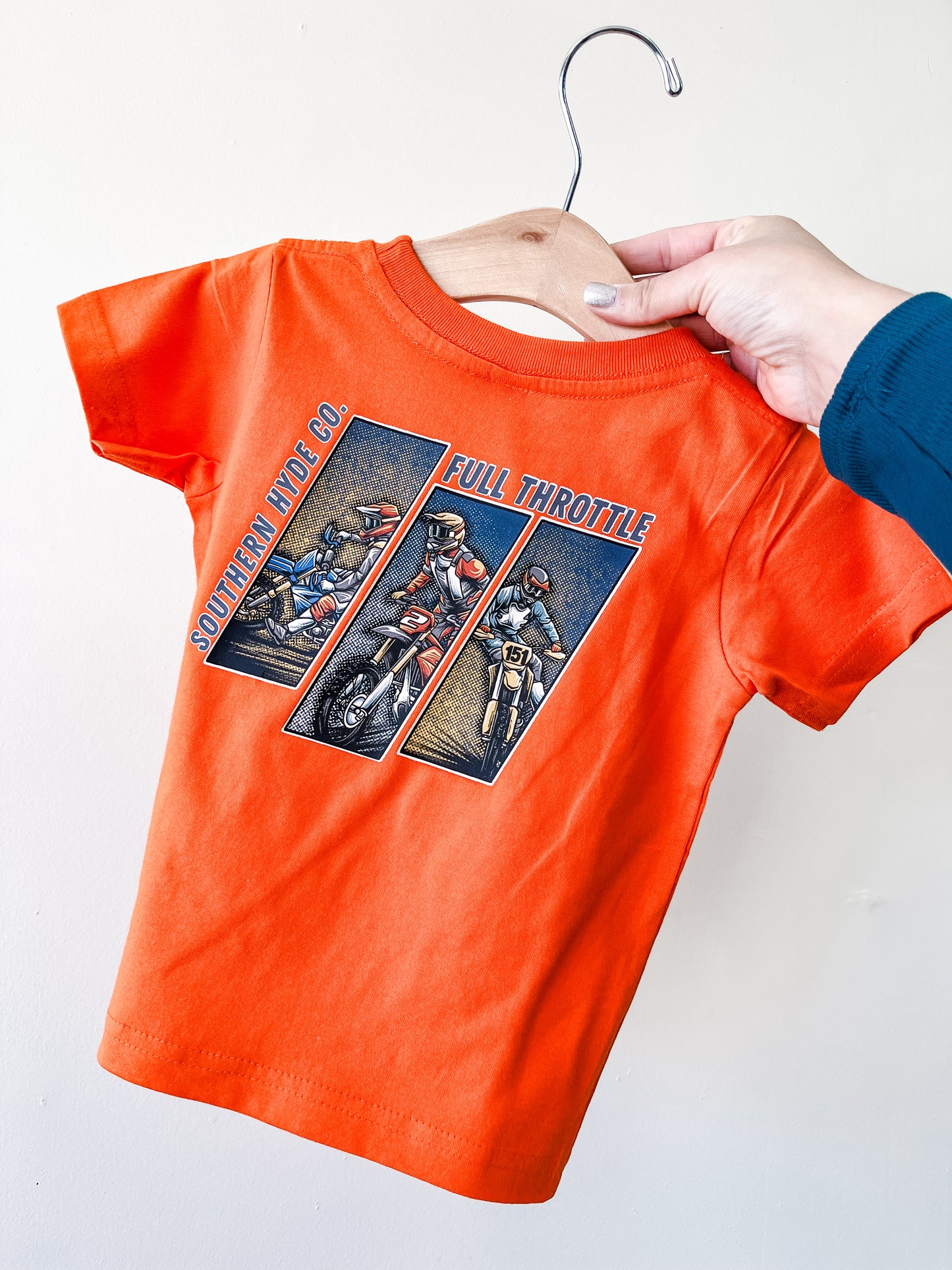 A hand holds up the Full Throttle Moto T-Shirt for kids by Southern Hyde Co—an orange tee with vintage motorcyclist illustrations and “Southern Hyde Co” plus “Full Throttle” printed on the front, displayed against a plain background.