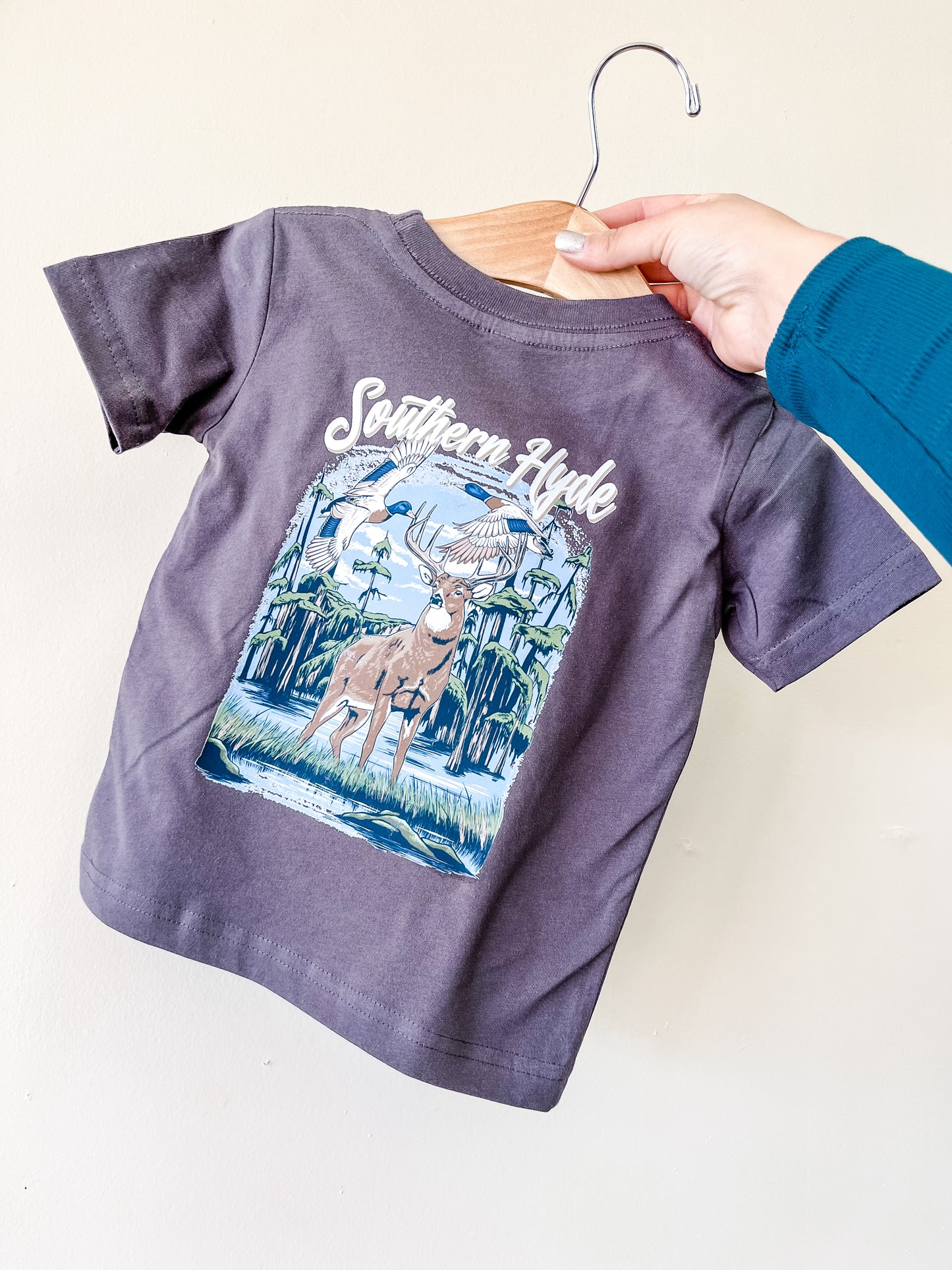 A hand holds a hanger with a gray children’s Hunter's Paradise T-Shirt by Southern Hyde Co, featuring an illustrated deer, two birds, trees, and “Southern Hyde” on the front.