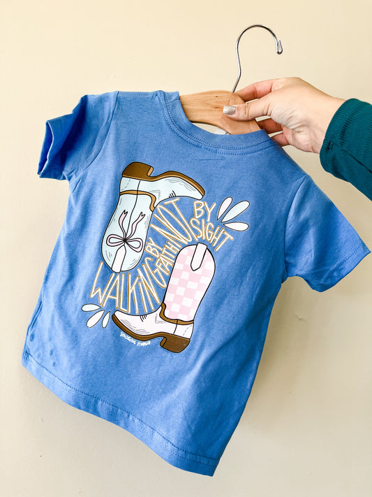 A hand holds Southern Hyde Co’s Walk by Faith T-Shirt, a blue children’s tee with illustrated boots and "Walking by faith, not by sight" in playful fonts on a wooden hanger.